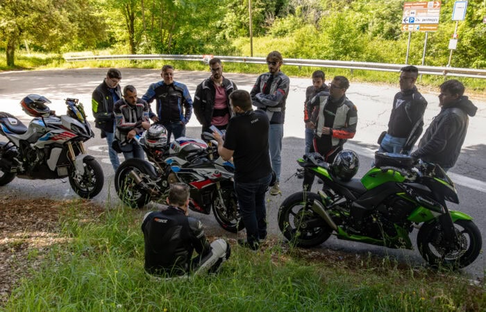 Gruppo di motociclisti che ascolta istruzioni con moto sportive e da turismo in un'area verde.